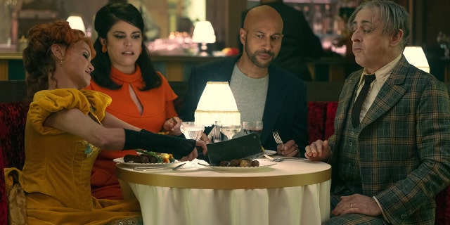 The cast of "Schmigadoon!" including Kristin Chenoweth in yellow, Cecily Strong in a bright orange, Keegan-Michael Key in a grey shirt and navy blazer and Alan Cumming in a green plaid suit set sit around a table in a photo from the second season