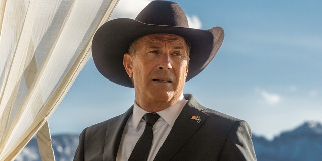 Kevin Costner in a black suit and black tie looks off in the distance wearing a cowboy hat for 'Yellowstone' photo as John Dutton