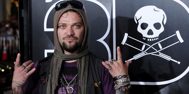 Bam Margera flashes his hands on the red carpet with sunglasses on his head and a scarf covering his head in a purple shirt