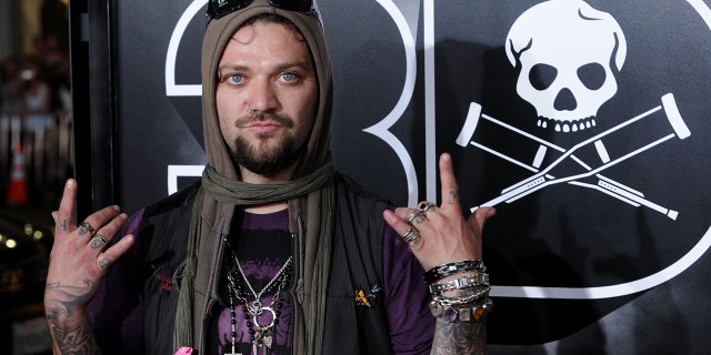 Bam Margera flashes his hands on the red carpet with sunglasses on his head and a scarf covering his head in a purple shirt