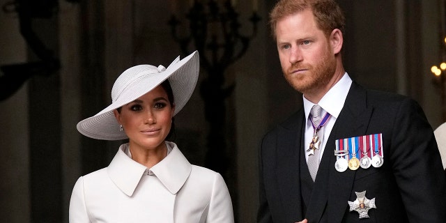 Meghan Markle wearing a white coatdress with a matching hat next to Prince Harry in a suit with his medals