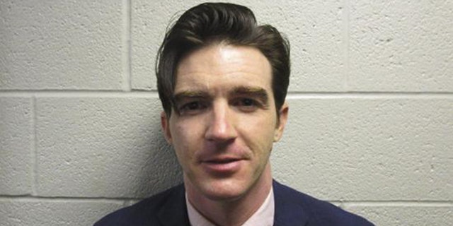 Drake Bell in a suit and tie in front of a white wall mugshot photo