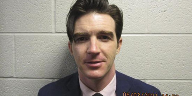 Drake Bell in a suit and tie in front of a white wall mugshot photo