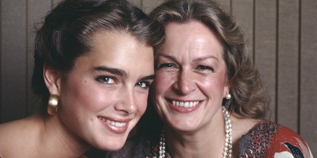 Brooke Shields in a colorful top and large gold earrings smiles next to her mother Teri Shields wearing a pearl necklace and earrings in a photo from 1981