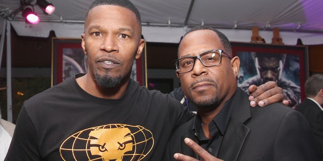 Jamie Foxx in a black shirt with a yellow graphic poses for a picture with Martin Lawrence, also wearing a black shirt doing a peace sign