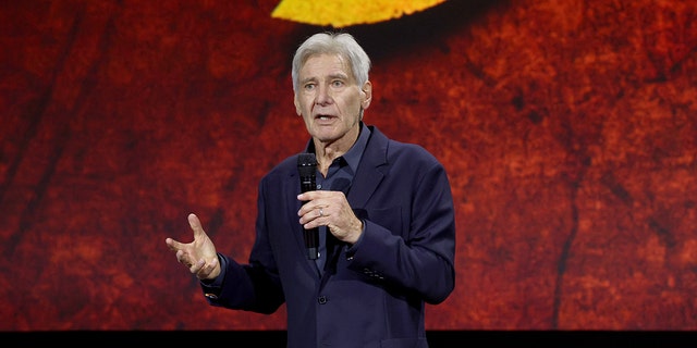 Harrison Ford wearing a dark blue shirt and blazer speaks into a microphone and holds his right hand out while on stage