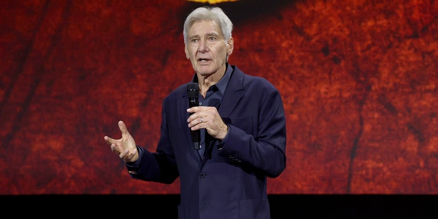 Harrison Ford wearing a dark blue shirt and blazer speaks into a microphone and holds his right hand out while on stage