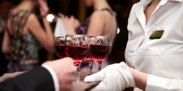 Close-up picture of waiter's hands wearing white gloves holding a tray with red wine, serving alcohol drinks. Catering service at special occasion, event. Hospitality industry concept.