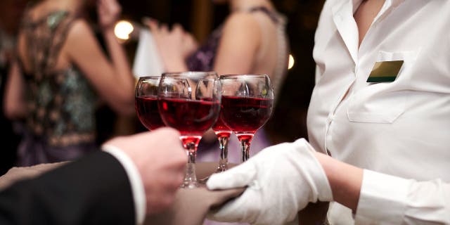 Close-up picture of waiter's hands wearing white gloves holding a tray with red wine, serving alcohol drinks. Catering service at special occasion, event. Hospitality industry concept.