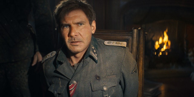 A still frame of Harrison Ford as Indiana Jones in a dirty blue jacket looking up and out ominously, his face is altered by AI