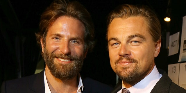 Bradley Cooper with a scruffy beard in a jacket and white shirt smiles next to Leonardo DiCaprio in a suit and tie and white shirt