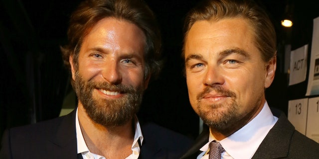 Bradley Cooper with a scruffy beard in a jacket and white shirt smiles next to Leonardo DiCaprio in a suit and tie and white shirt