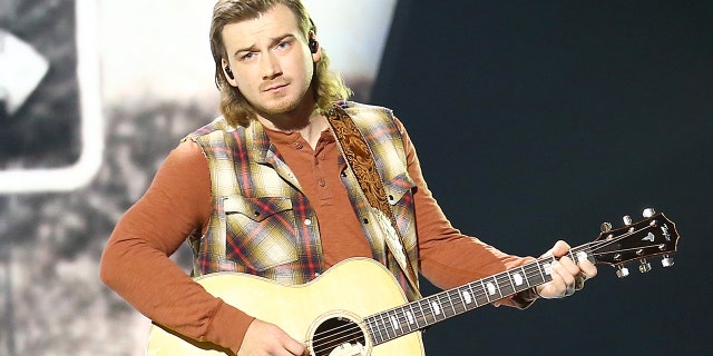 Morgan Wallen strums a guitar on stage while wearing plaid vest and jeans