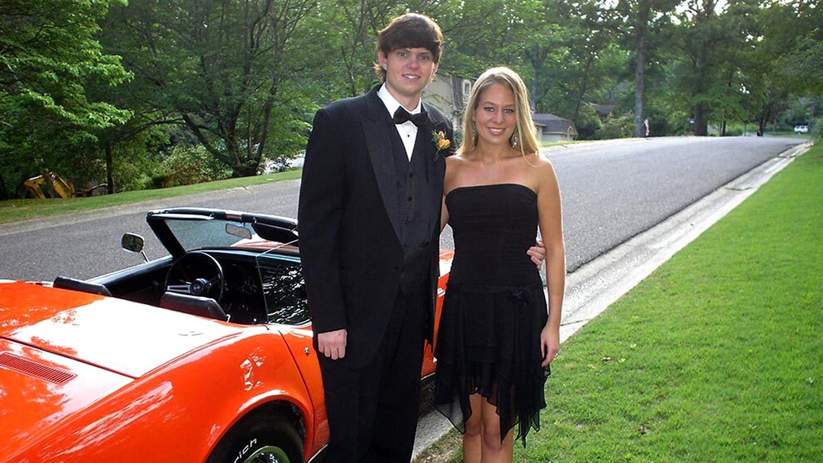Henry in a tuxedo, Natalee in a short black dress, the pair stand in front of a convertible red muscle car