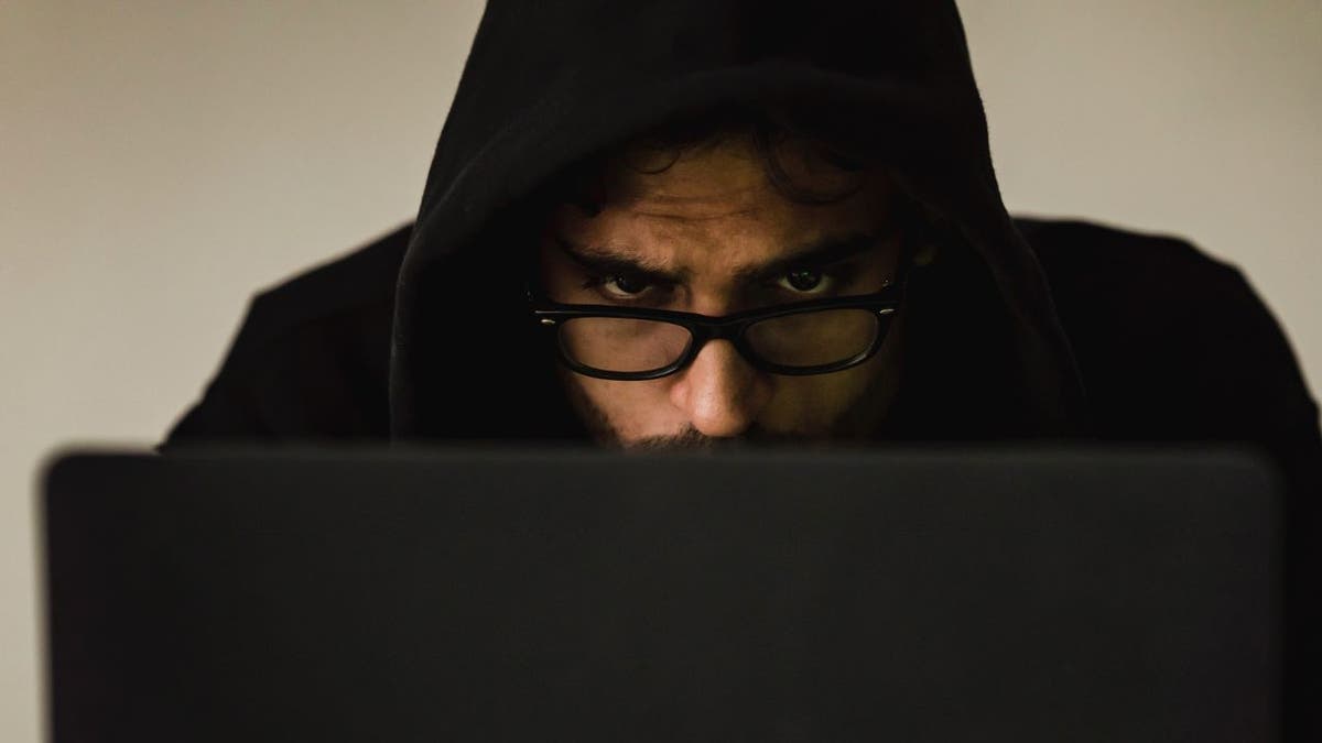 Man in black hoodie with glasses hovering over black laptop