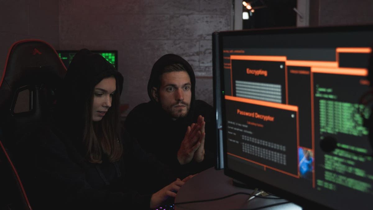 Girl and guy in black hoodies sitting next to computer screens hacking 