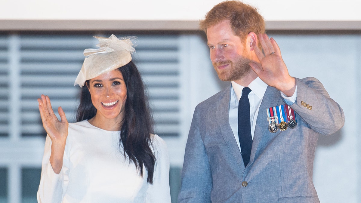 Meghan Markle wearing a creme dress and a matching hat next to Prince Harry in a grey suit and black skinny tie