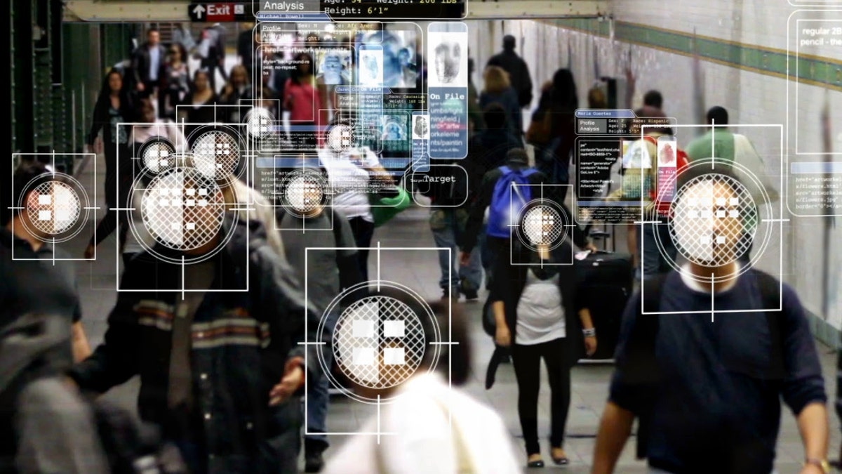 A computer scans a crowded subway tunnel, using facial recognition to show personal data in a floating display that follows the individual. Each display has a photo of the person, a unique fingerprint, and basic information like name, race, age, etc. There are small blocks of text which have scrolling code excerpts.