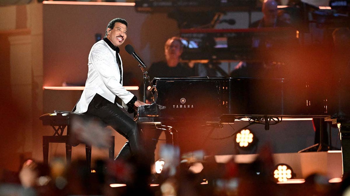 Lionel Richie wears a white blazer with sparkling rhinestones while sitting behind a piano at coronation concert