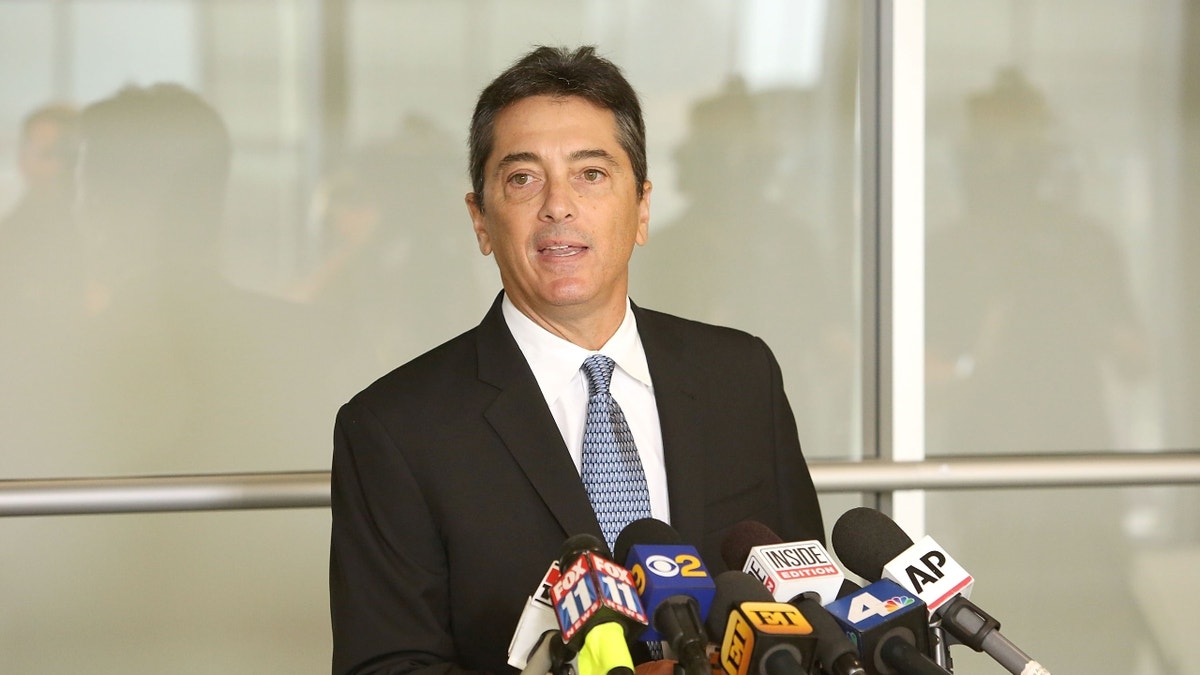 Scott Baio behind podium in a black suit, white button-down and blue tie