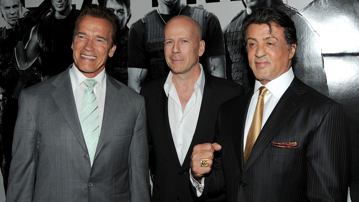 Arnold Schwarzenegger in a grey suit and green tie stands next to Bruce Willis with a white shirt and black jacket next to Sylvester Stallone flashing a fist in a black suit and gold tie at the premiere of "The Expendables"