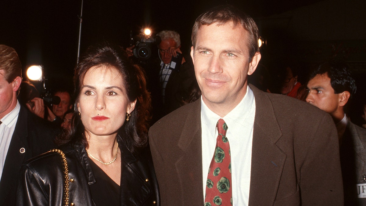 Kevin Costner em um terno marrom olha ao longe com sua esposa Cindy de preto na Califórnia