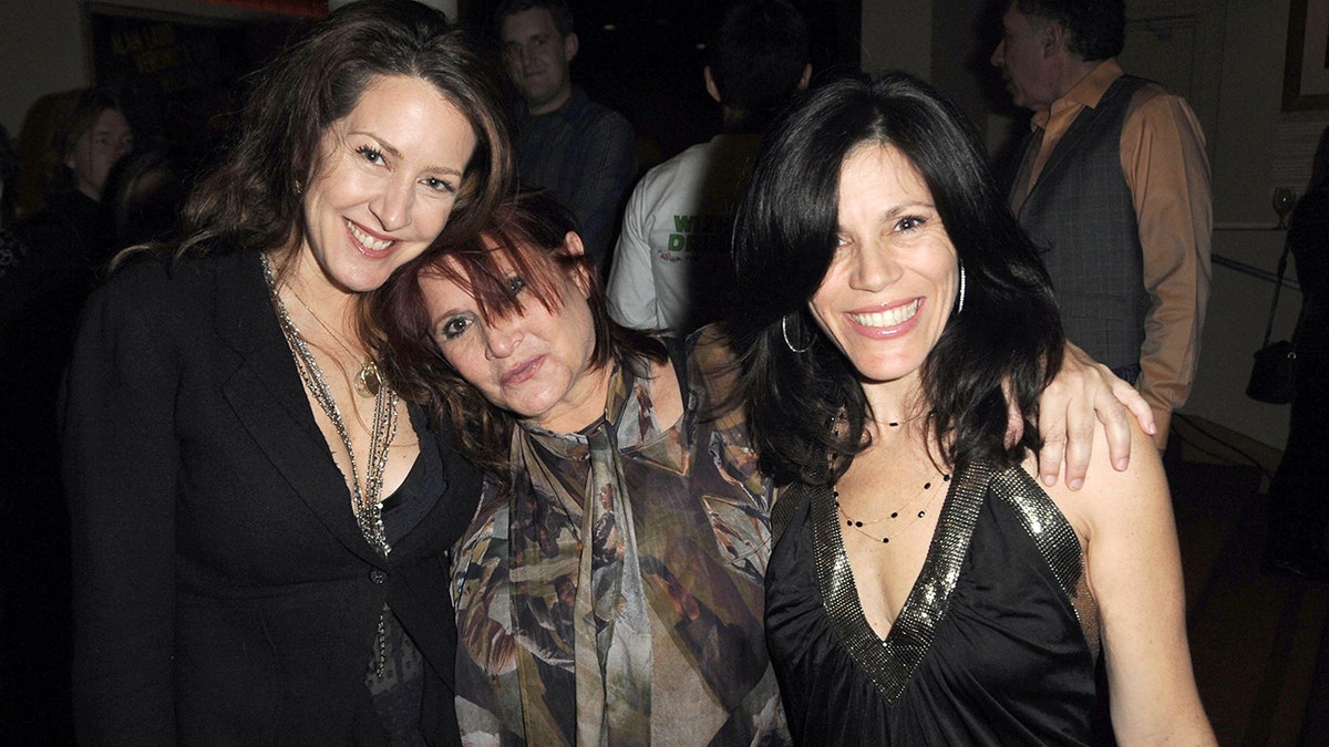 Joely Fisher in black and a long chain necklace is embraced by Carrie Fisher in a patterned top leaning her head on Joely's shoulder, next to Tricia Leigh Fisher also in a black v-cut top