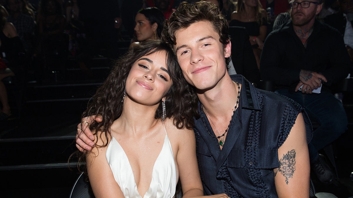 Camila Cabello in a white dress smiles as Shawn Mendes wraps hi arm around her