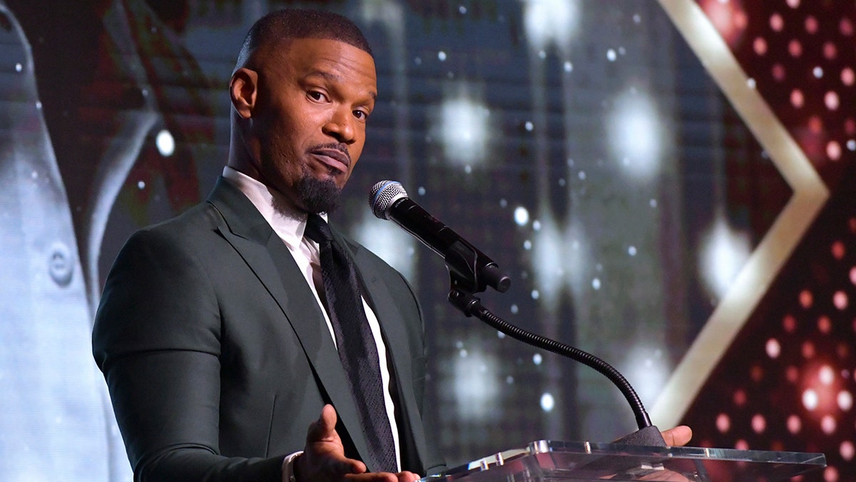 Jamie Foxx looks at the camera behind the podium at the AAFCA Awards wearing a dark suit and black tie