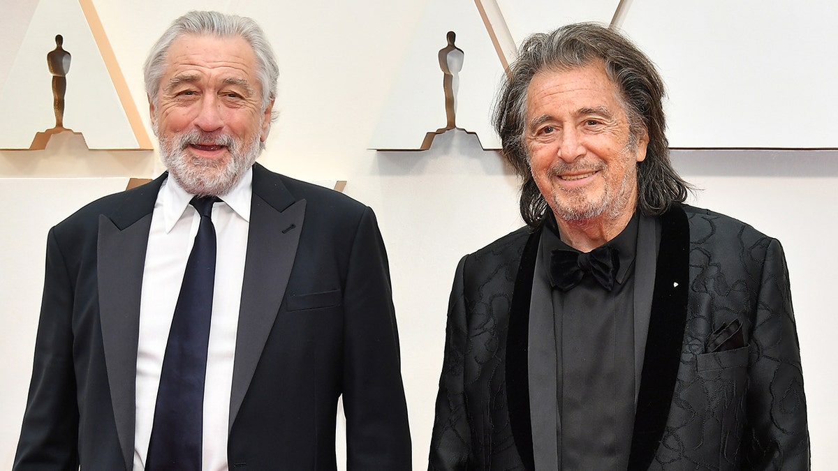 Robert De Niro smiles in a black suit and tie next to Al Pacino in a black suit at the Academy Awards in 2020