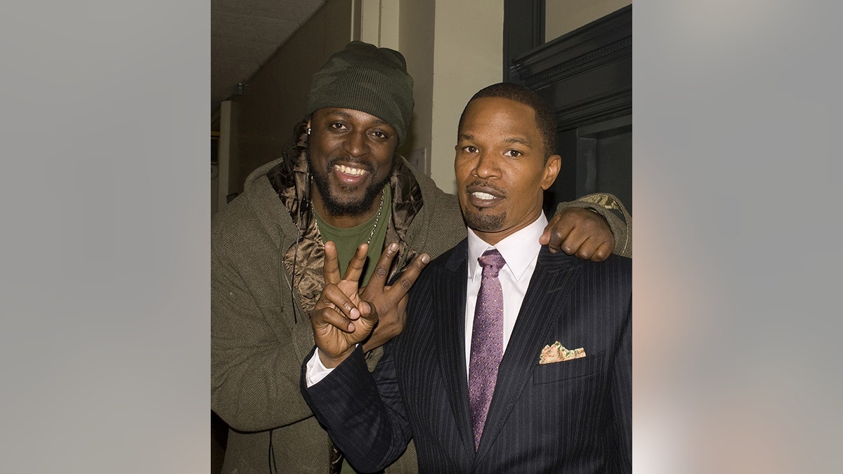 Jamie Foxx in a suit and purple tie holds up the peace sign with Charlie Mack, also holding a peace sign in a green hoodie and hat