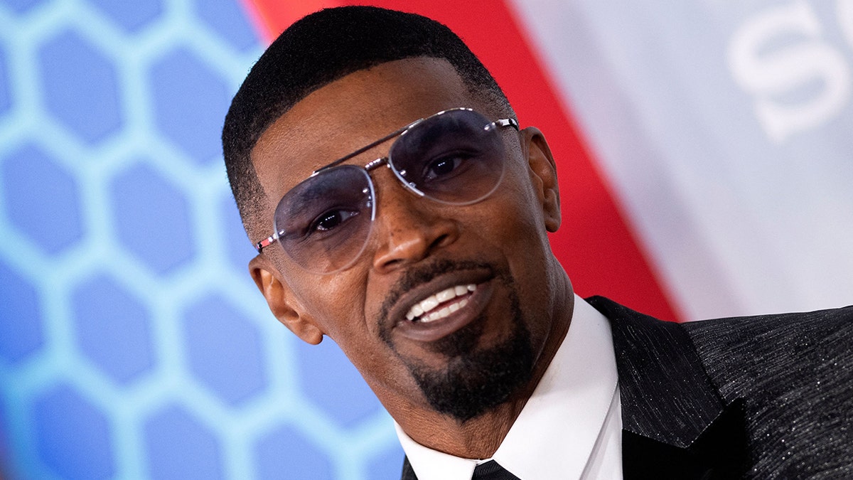 Jamie Foxx smiles on the red carpet in a suit, white shirt and tie with tinted sunglasses