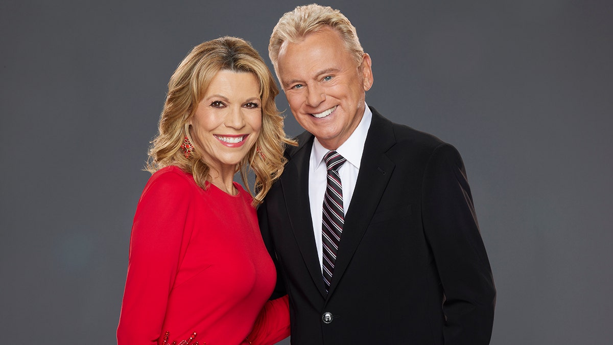 Vanna White in a red dress smiles in a promotional photo for "Celebrity Wheel of Fortune" with Pat Sajak in a black suit and striped tie
