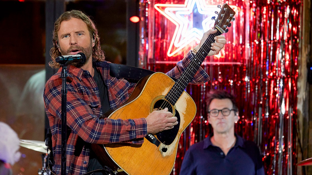 Dierks Bentley in a red plaid holds up a guitar and strums in front of a microphone