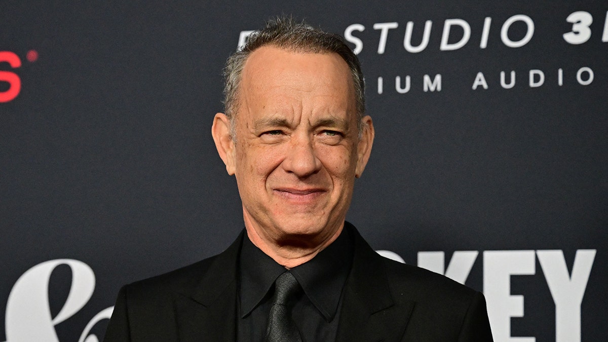 Tom Hanks smiles with his mouth closed wearing a black suit at the LA Convention Center in LA