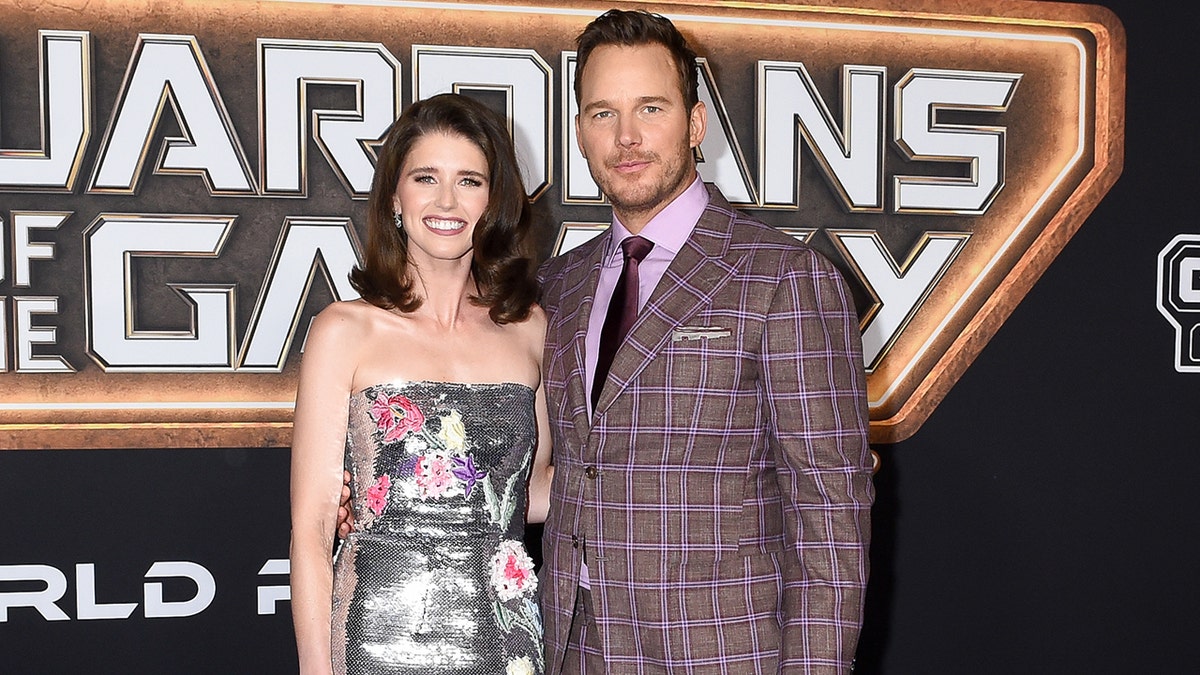 Katherine Schwarzenegger Pratt in a silver dress with pink detail smiles on the red carpet with husband Chris Pratt in a purple/brown checkered suit, lilac button-down and dark tie