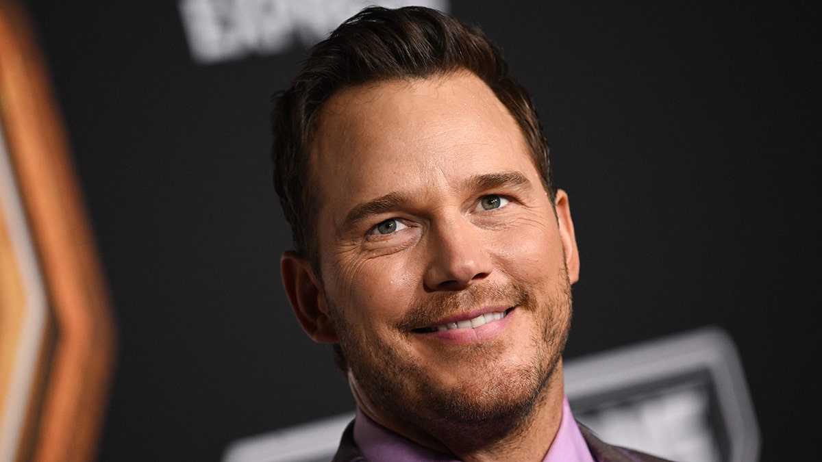 Chris Pratt smiles and looks upward in a bright lilac shirt and suit on the red carpet