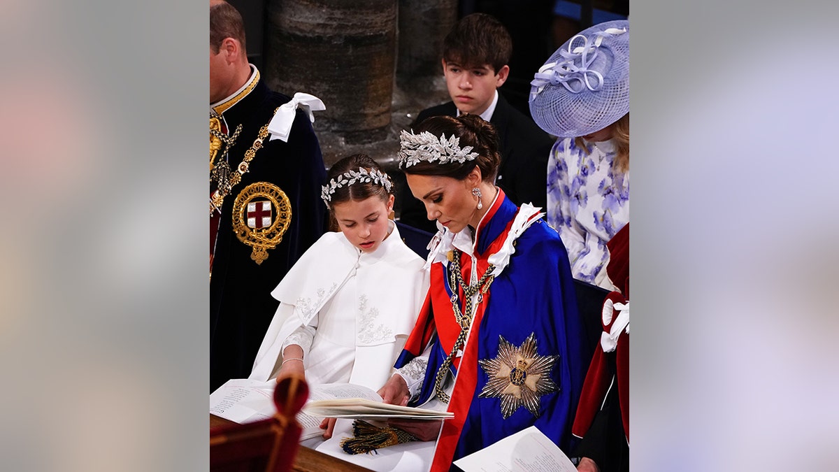 Inside Westminster Abbey, Princess Charlotte in white and a leaf tiara looks at a reading with her mother Catherine, Princess of Wales, in a blue robe with red ribbon and a matching tiara