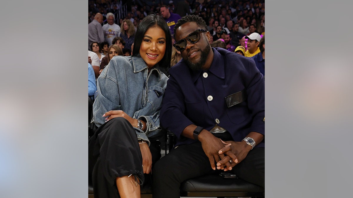 Eniko Hart in a denim jacket sits next to husband Kevin Hart wearing black sunglasses inside at the Lakers game