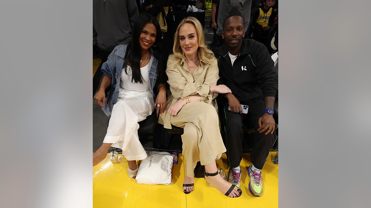 Nia Long in white and a denim jacket poses next to Adele in a tan jumpsuit, holding the arm of boyfriend Rich Paul, in black, sitting courtside