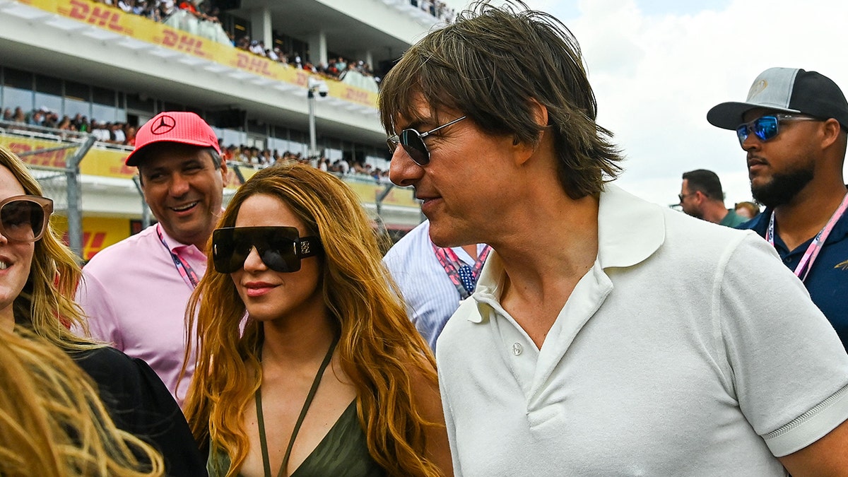 Shakira in a black tank top with two strings in the center around her neck and massive glasses walks alongside Tom Cruise in a white polo shirt at the Grand Prix in Miami