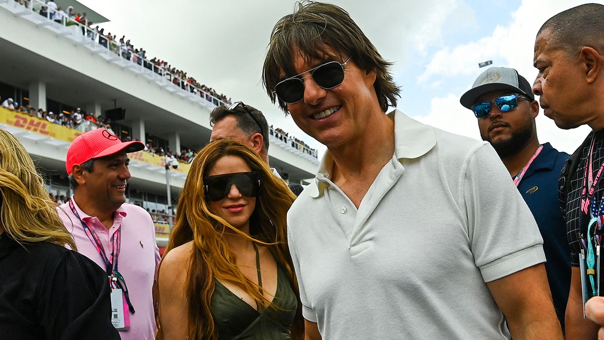 Shakira in a black tank top with two strings in the center around her neck and massive glasses soft smiles for a picture with Tom Cruise in a white polo shirt at the Grand Prix