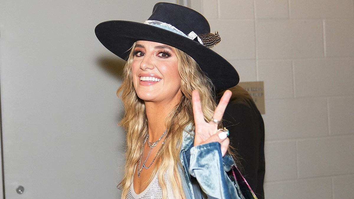 Lainey Wilson dá às câmeras um sinal de paz enquanto entra no Country Music Awards, usando um chapéu de aba preta