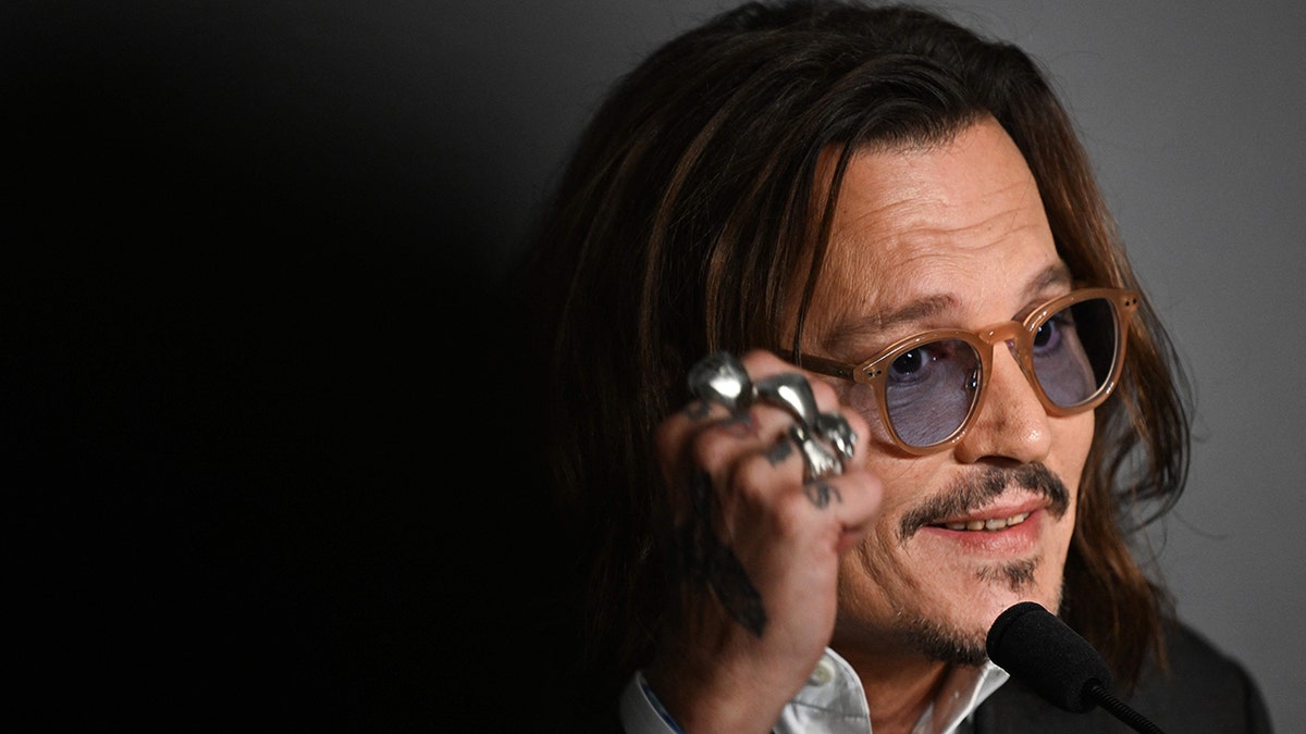 Johnny Depp wears brown rimmed glasses with blue lenses and speaks into a microphone while doing press at Cannes