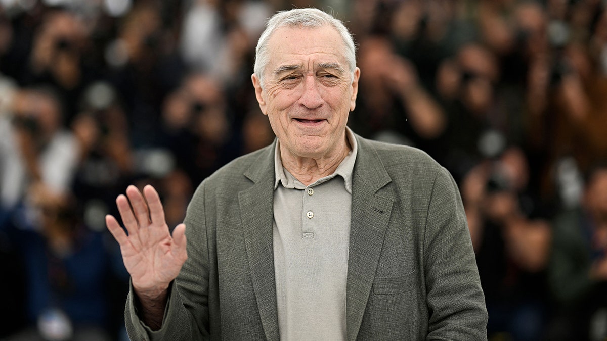 Robert De Niro in a light grey shirt and darker grey jacket waves to the crowd in Cannes