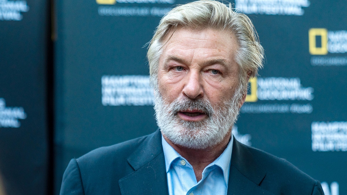 Alec Baldwin with a beard looks off camera on the red carpet, wearing a blue shirt and blazer