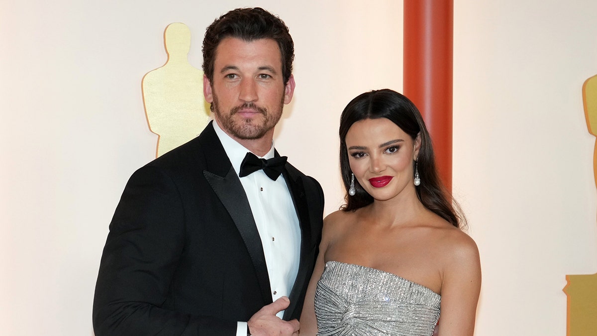 Miles Teller in a tuxedo smiles on the carpet with wife Keleigh in a silver gown at the Oscars