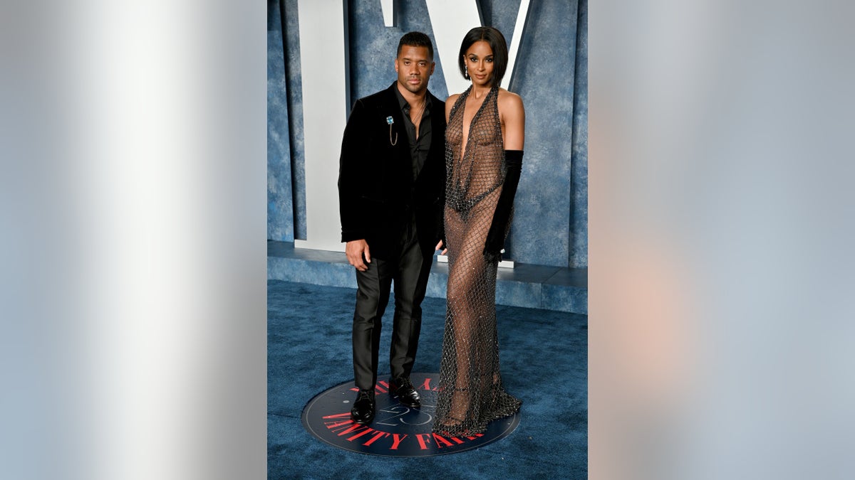 Russell Wilson in a black outfit at the Vanity Fair Oscar Party holds onto Ciara in a mesh/fishnet dress
