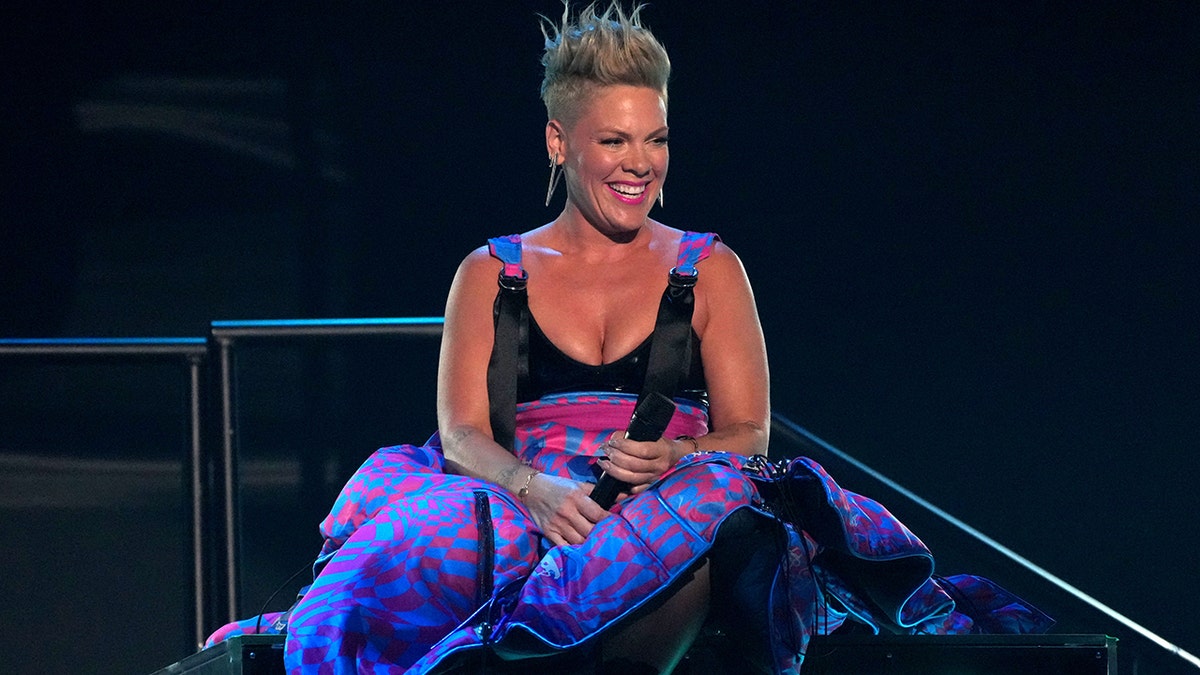 Pink sits on stage in a black top/printed bottom dress and smiles at the iHeartRadio Music Awards