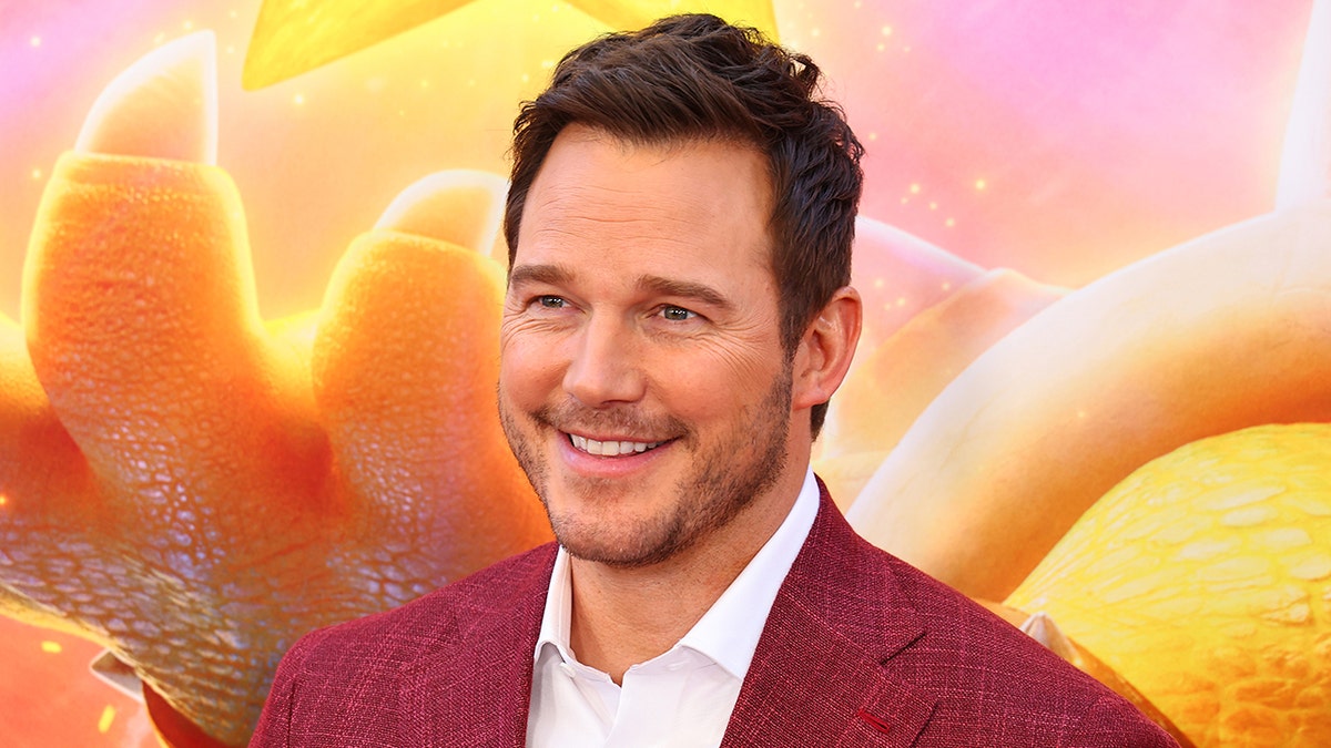 Chris Pratt wearing a white button down and red jacket smiles on the red carpet